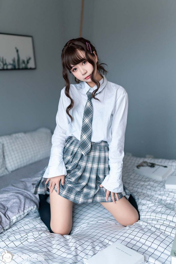 一位可爱的文艺学妹，身穿灰色格子裙JK制服和黑色长袜，坐在床上，手里捧着一本书。